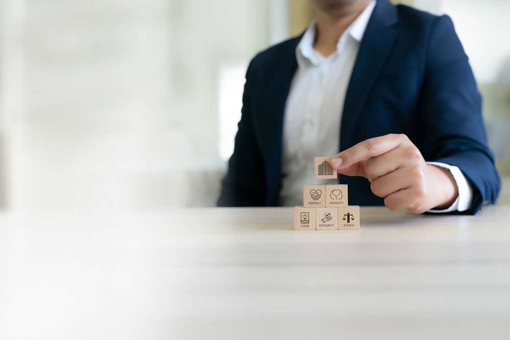 Homme en costume empilant des blocs de bois représentant les icônes du code de conduite : code, intégrité, honnêteté, respect et éthique, les fondements d'un code de conduite d'entreprise solide.