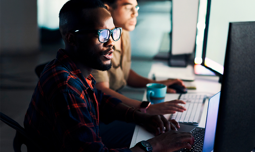 Tech professionals working on computers in a dim workspace, focused on code and digital analysis