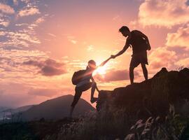 One person helps another up a mountain with a sunset background