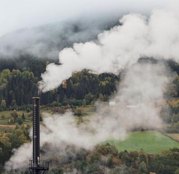 Factory chimney emitting smoke against a forest backdrop