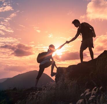 One person helps another up a mountain with a sunset background