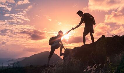 One person helps another up a mountain with a sunset background